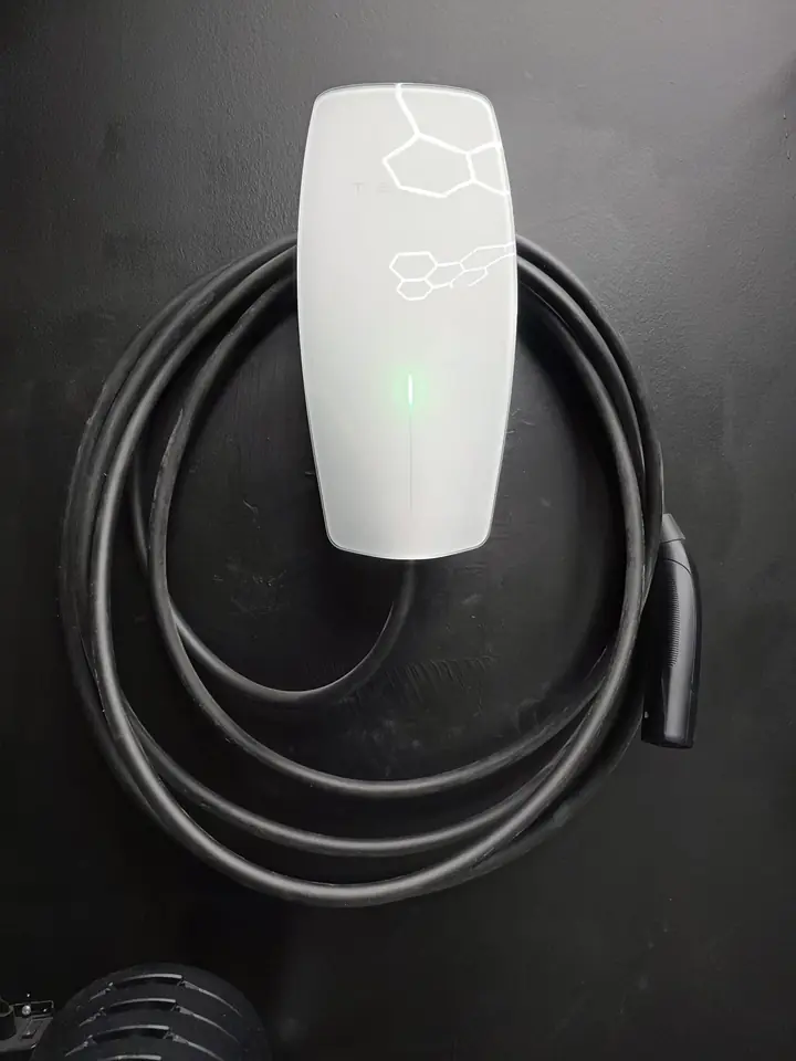 White Tesla Wall Connector on a dark garage wall, cable coiled on the holster, status light on