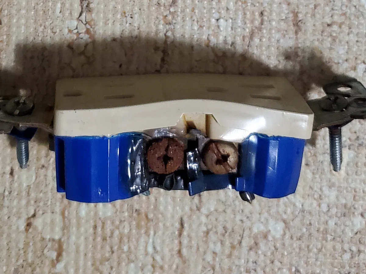 The outlet pulled from behind the couch, its screws dark from heat and the plastic around them melted