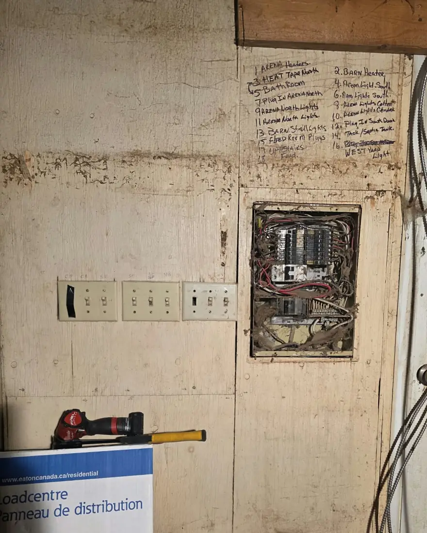 Old panel recessed into a plywood wall at a horse ranch, with the circuit list written in marker above it and three switches beside it