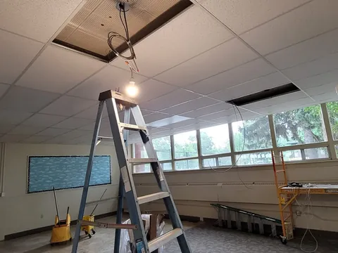A commercial room with ceiling tiles open and the old fixtures coming out