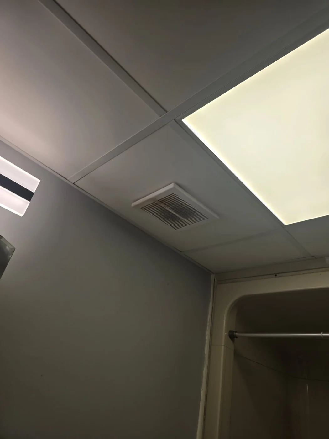 Bathroom drop ceiling before the work, with a small white slotted exhaust fan grille beside a flat 2 ft by 2 ft LED panel.